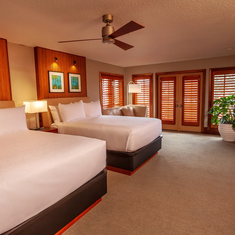 Double Queen Bedroom - Villa 2 An image of a double queen bedroom in an Evermore Orlando Resort Villa. The large room features two plush queen beds, multiple lamps, a large ceiling fan, a walk mounted TV above a work desk and a set of doors that lead outside to an exterior patio.